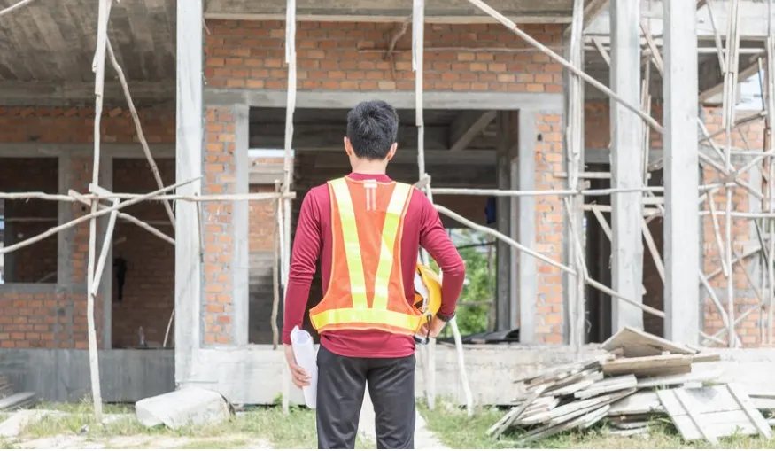 person-wearing-a-construction-vest-with-his-back-turned-looking-towards-a-house-under-construction